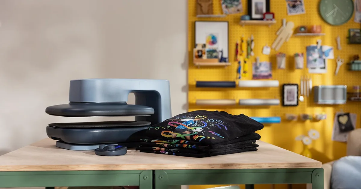 Cricut AutoPress 2 automatic heat press on a workbench beside a stack of folded black DTF-printed T-shirts