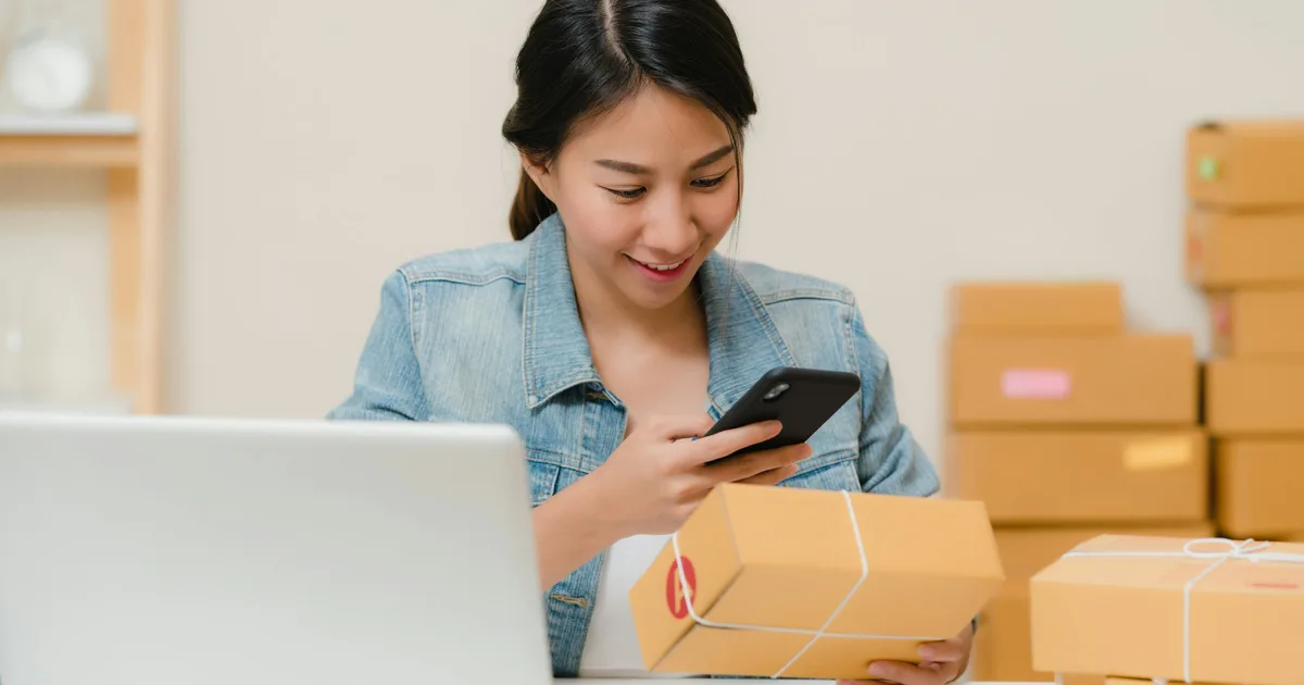 Small business owner checking her phone while holding a kraft shipping box, with stacked boxes and a laptop on the desk