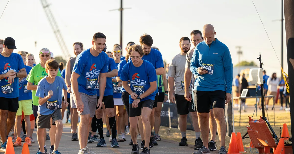 Runners at a community fun run start area wearing matching blue event t-shirts with race bibs