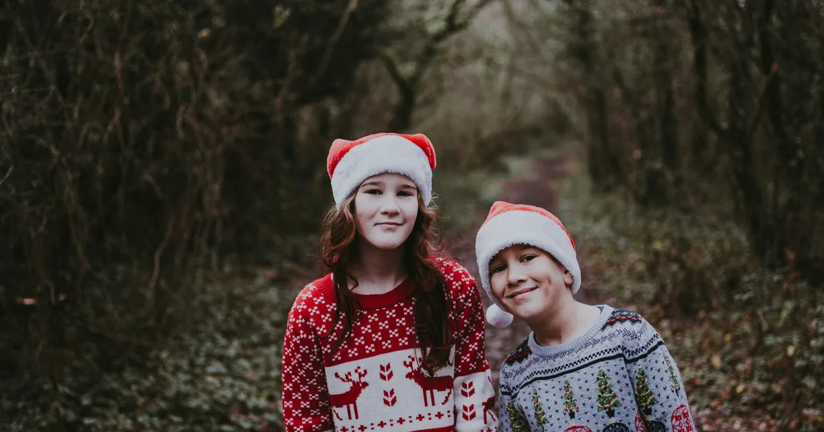 Two children in Santa hats wearing red and white reindeer-pattern Christmas sweaters outdoors