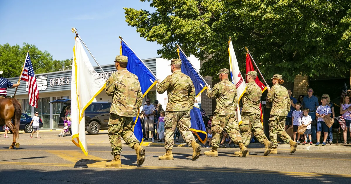 Uniformed U.S. color guard carrying the American flag and military flags in a hometown parade as families watch from the sidewalk